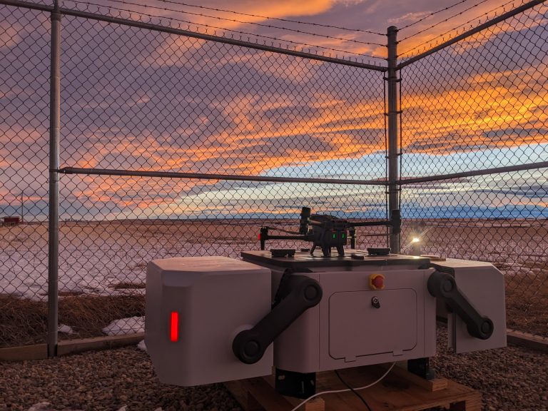 DJI drone docking station set behind fence. Background is facility site with a sunset